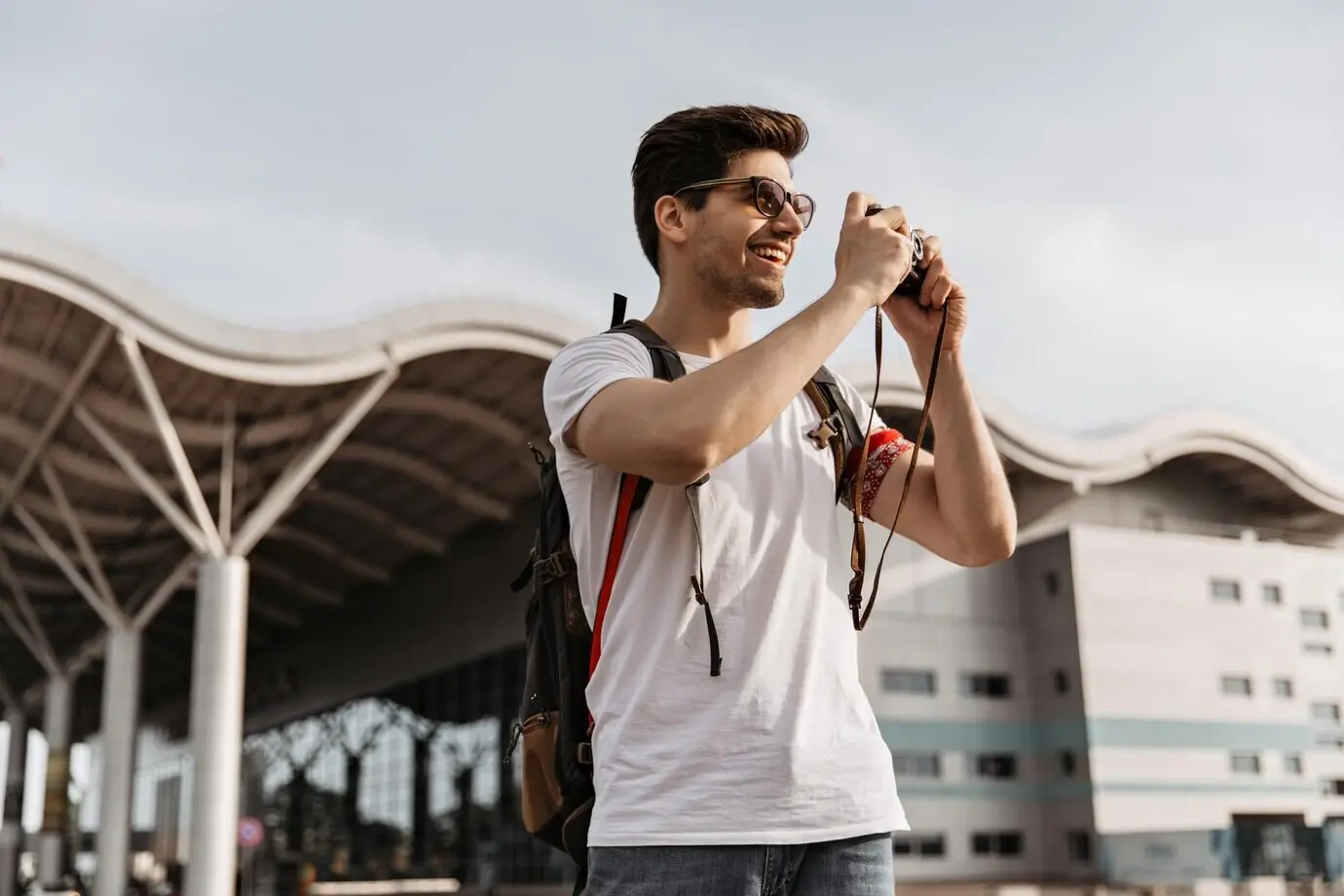 Glücklicher Mann in Jeans und weißem T-Shirt fotografiert draußen in der Nähe eines Flughafens. Cooler brünetter Mann mit Sonnenbrille hält einen Rucksack und eine Retro-Kamera.